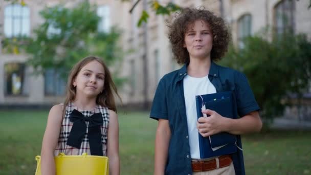 屋外に立ってカメラを見て手を振って話す白人の男の子と女の子の肖像画 フロントビューメディアショットの自信のある学童ブロガーポージング広告教育 — ストック動画