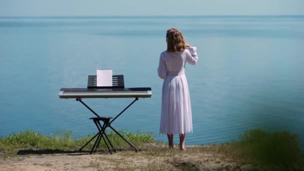 戻る青い川の波の背景にピアノ シンセサイザーに立って裸足の10代の少女を表示します 広いショット思慮深い白人青年音楽家離れて見て夢見ている スローモーション — ストック動画