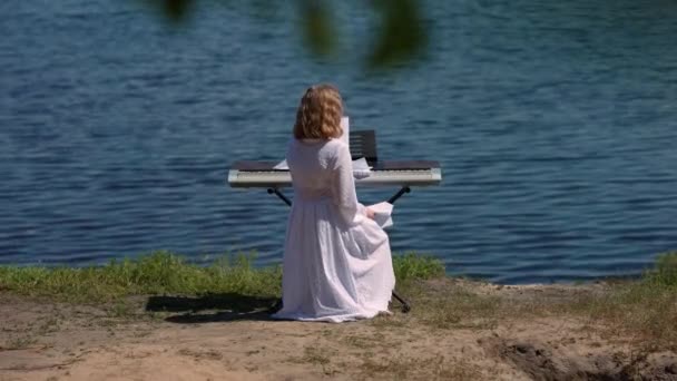 先頭へ戻る10代の白人の少女は 晴れた春の夏の日に青い川の水の背景にピアノ シンセサイザーに座っています 自信を持って10代の音楽家が紙飛行機で海岸に歩いて立ち上がる — ストック動画