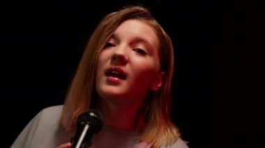 Sahnede şarkı söyleyen yetenekli kendine güvenen genç kız mikrofonda aşağı yukarı hareket ediyor. Güzel, beyaz tenli ergen şarkıcı sahne ışıklarıyla karanlıkta şarkı söylüyor.