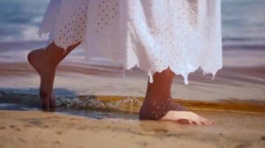 Yakın plan kadın ayakları sahil manzaralı altın kumda yürüyor. Tanımlanamayan beyaz bir kızın gün ışığında suyun içinde gezinirken görüntüsü. Doğa ve eğlence kavramı