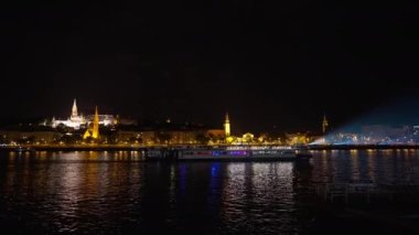 Şehrin arka planında, gece nehri üzerinde geniş açılı su taksisi. Kasaba ışıklarının arkasında, karanlıkta yolcularla dolu bir gemi.