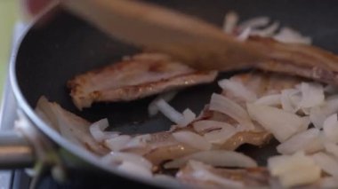 Pastırmayla soğanı, mutfakta kızartma tavasında yakın plan karıştırın. Çatallı beyaz bir erkek, evde lezzetli yemekler pişiriyor.