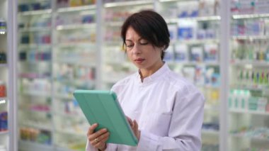 Konsantre kadın, eczanede durup yavaş çekimde gülümseyen kameraya bakarak dijital tablet dokunmatik ekranı kaydırıyor. Eczanede poz veren kendine güvenen zeki bir çalışanın portresi.