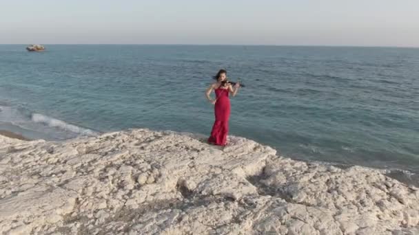 地中海沿岸に立つバイオリンを演奏赤いドレスでコーカサスの豪華な女性のドローンビューをズームアウトします 崖の上で屋外で実行するワイドショット才能ある自信アーティスト — ストック動画