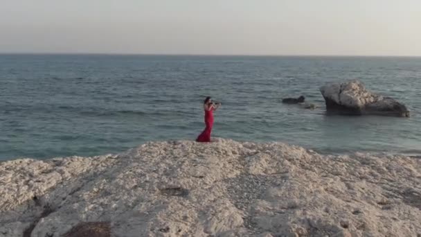 地中海の波の背景に崖の上に立ってバイオリンを演奏吸収ミュージシャンにドローンビューズーム ワイドショット自信を持って白人才能のある女性屋外でキプロス海岸 — ストック動画