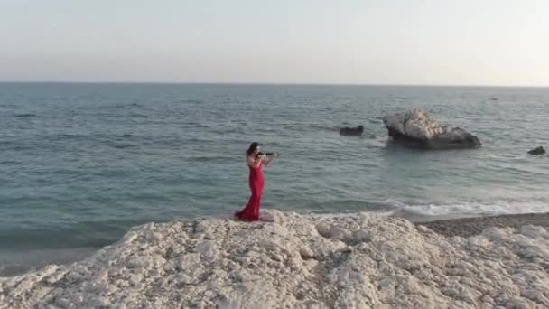 海岸の崖の上で実行される赤いドレスで才能のある女性に空中ズーム 地中海の海岸で太陽の下で風の強い日にバイオリンを演奏才能のある白人音楽家 — ストック動画