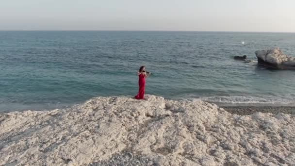 海の崖の上に立つバイオリンを演奏赤いドレスで自信に満ちた才能の白人女性の肖像画屋外 キプロスの地中海で屋外音楽を楽しむ広いショット豪華な美しい女性 — ストック動画