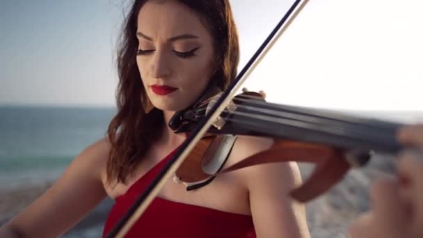 地中海の海岸で太陽の光の下で立って音を楽しんでバイオリンを演奏する深刻な女性ミュージシャンの肖像画 日没時に海の海岸に集中才能のある白人女性 — ストック動画
