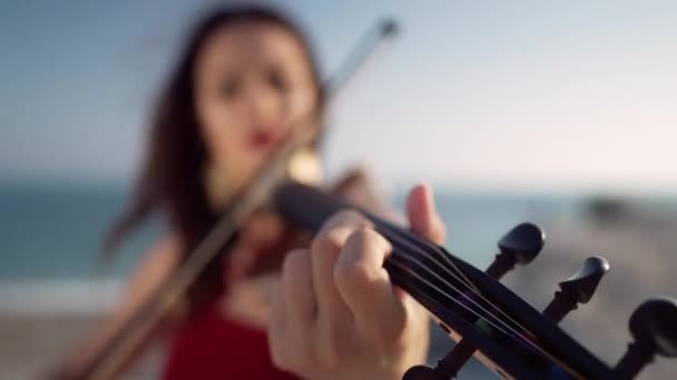 背景に弓で遊んでいるぼやけた白人ミュージシャンとバイオリンのフリットボード上のクローズアップ女性の指 海の海岸で音楽を演奏する認識できない才能のある女性 — ストック動画