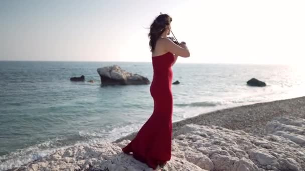 サイドビュー才能のある女性が海の波が日没時に海岸に圧延としてバイオリンを演奏 広いショットコーカサスの自信のあるミュージシャンで 地中海のビーチで日没時に屋外で演奏する赤いドレス — ストック動画
