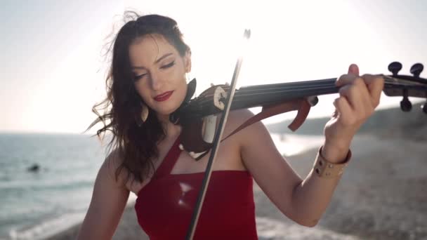 日の出の太陽の下でインスピレーションを受けた女性の肖像は スローモーションでバイオリンを演奏します フロントビュー地中海の海岸で音楽を演奏ポーズ自信を持って白人の才能のあるミュージシャン — ストック動画