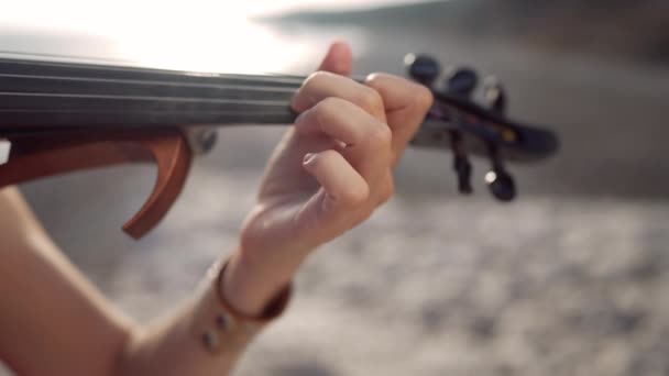 ゆっくりとした動きの中 女性の手がバイオリンの板の上に弦に触れている 太陽の下で地中海のビーチで日の出に音楽を演奏認識できない白人女性 — ストック動画