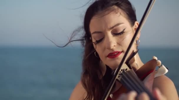 日没時に海の海岸でバイオリンを弾いているカメラを見て自信のある女性のクローズアップ肖像画 地中海沿岸の弦楽器で音楽を演奏する才能豊かな白人アーティスト — ストック動画