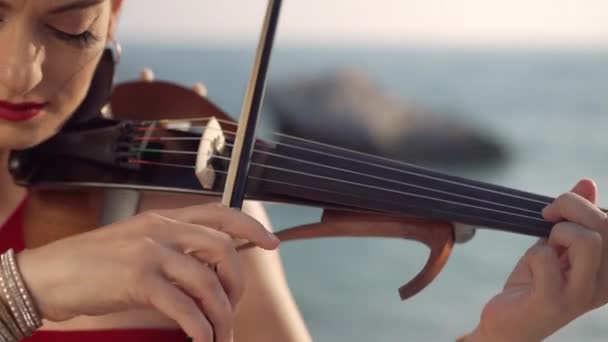 日光の屋外で音楽を演奏白人女性の肩の上にクローズアップバイオリン 日没時に地中海沿岸の海の海岸で自由を楽しんで自信を持って才能のある女性 — ストック動画