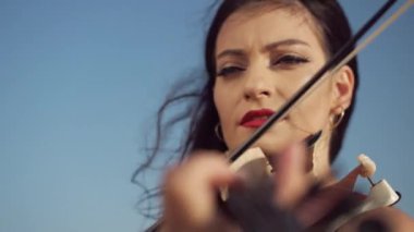 Yakın plan yetenekli kadın bulutsuz mavi gökyüzünün arka planında keman çalıyor. Kafkasyalı kadının kendine güvenen portresi. Kameraya bakıyor. Açık havada gün ışığında müzik yapıyor.