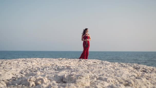 海の崖の上に立つバイオリンと赤のドレスでワイドショットスリム豪華な女性が離れて見て フロントビュー地中海沿岸の日差しの中で美しいコーカサスの才能あるミュージシャン スローモーション — ストック動画