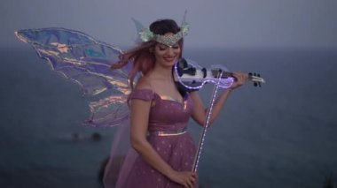 Yetenekli, gülümseyen kadın elinde keman ve okla okyanus kayalığında karanlıkta duran ışığı tutuyor. Peri kostümü giymiş, enstrümanlı beyaz müzisyenin orta boy portresi.