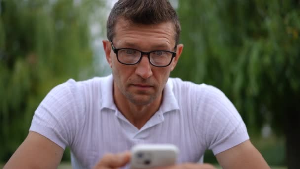 フロントビューの男は電話アプリで驚きと困った表情読み取りメッセージを作っています 春の夏の公園で屋外スマートフォンを持つストレス白人男性の肖像画 スローモーション — ストック動画