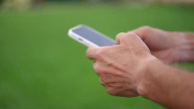 Yakından beyaz erkek eller akıllı telefon dokunmatik ekranında yavaş çekimde yazı yazıyor. Çevrimiçi açık havada yeşil çimenlerin arkasında sosyal medyayı kaydıran tanınmamış bir adam mesajı gönderiyor.