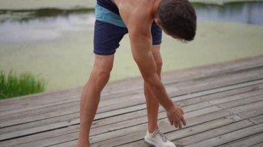 Canlı kamera sporcuyu takip ediyor. Ayaklarını uzatarak ayağa kalkıyor ve ağır çekimde kollarını kaldırıyor. Motive olmuş Kafkasyalı yetişkin adam yaz parkında dışarıda çalışıyor.