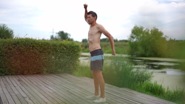 春天的夏季公园里 一个体格健康的高加索人手拉手站在湖边 广博的侧视自信的有动力的运动员在室外慢动作热身训练 — 图库视频影像