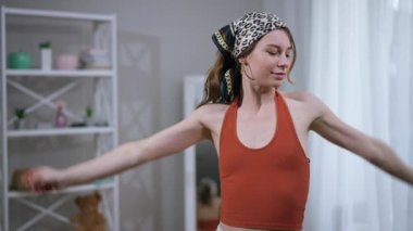 Sıska genç bir kadın dans ediyor aynaya doğru yürüyor başörtüsünü çıkarıyor ve kameraya bakarak geri dönüyor. İçerideki modadan hoşlanan kendine güvenen beyaz kadın portresi.