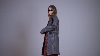 Palto ve güneş gözlükleriyle gri arka planda dönen genç model gülümsüyor. Kendinden emin beyaz kadın portresi.