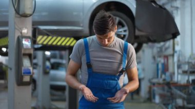 Kirli elleri olan, üniformalı yakışıklı genç adam araba servis istasyonunda dikilip ağır çekimde etrafa bakıyor. Beyaz tenli tamircinin orta boy portresi.