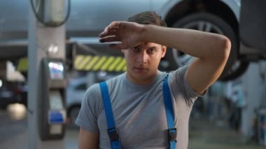 Alnındaki teri koluyla silen genç bir adam kameraya bakıyor. Kafkasyalı erkek oto tamircinin portresi. Servis istasyonunda yavaş çekimde poz veriyor.