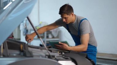 Dijital tabletli, İngiliz anahtarı silen alnıyla motor parçalarını beceren beyaz bir adam. Benzin istasyonunda bozuk araba tamir eden yakışıklı, konsantre beyaz tamircinin portresi.