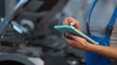 Garajdaki açık araba kaputunun arka planında erkek ellerinde dijital tablet var. Tanımlanamayan beyaz araba tamircisi servis istasyonunda uygulaması olan aracı kontrol ediyor.