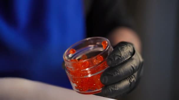 一个无法辨认的人用红色鲑鱼鱼子酱填满小玻璃瓶 男性白种人手戴手套 把美味的美味佳肴放在室内的集装箱里 — 图库视频影像