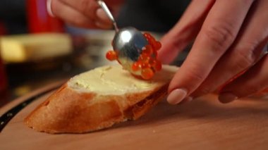 Tereyağlı ve kadın eli ile yakın plan lezzetli sandviç yavaş çekimde kırmızı somon havyarı yayıyor. Tanımlanamayan genç beyaz kadın içeride atıştırmalık hazırlıyor.