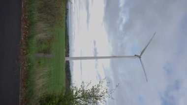 Yeşil çayır üzerinde bulutlu gökyüzünün arka planında dönen yel değirmeninin dikey videosu. Geniş açılı rüzgar türbini, açık havada yenilenebilir enerji üretiyor.