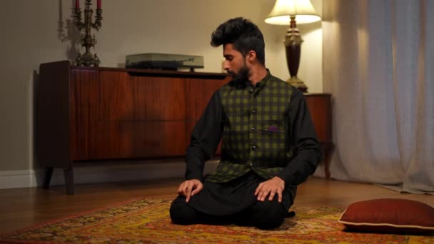 若いイスラム教徒の男性は 自宅のリビングルームでラマダーン月に祈る側に座っていました 広いショットの肖像画の深刻な濃縮中東男上の膝上のカーペット室内 — ストック動画