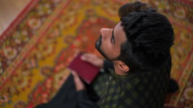 Yüksek açılı, Kuran 'lı genç Müslüman adamın Ramazan ayını kapalı bir mekanda namaz kılarken portresi. Geleneksel ayinler yapan kendinden emin Orta Doğulu inananların omuzlarından ateş etmek