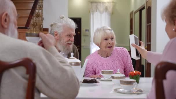 年配のカップルがお茶会で友人のカップルと座って賞賛の写真フレームを話しています 積極的な白人老人と女性楽しみますレジャーオン退職室内 — ストック動画