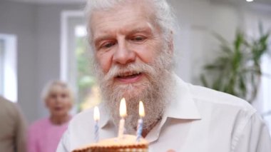 Sakallı mutlu son sınıf öğrencisinin portresi. Pastaya mumları üflerken gülümsüyor. Beyaz erkek emekli doğum gününü arka planda sevinçle alkışlayan bulanık arkadaşlarıyla kutluyor.