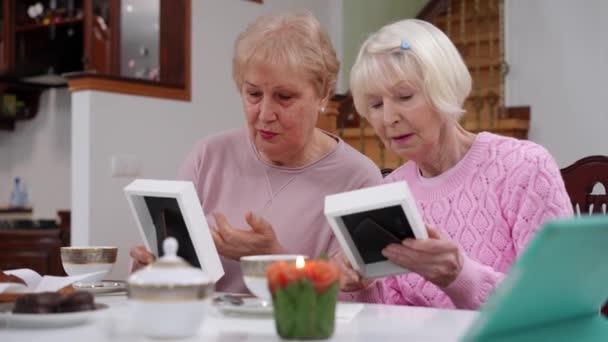 2人の年配の女性が居間のテーブルに座って古い写真を見て話していた 室内で出会う記憶を想起させる白人の退職者の肖像 — ストック動画