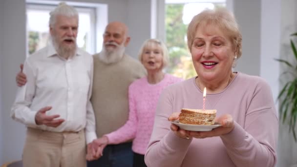 女性の退職者は喜びを拍手ぼやけた友人を振り返って誕生日に願い事を作るろうそくを吹いて笑顔 家の中で休日を祝う白人のシニア女性の肖像 — ストック動画
