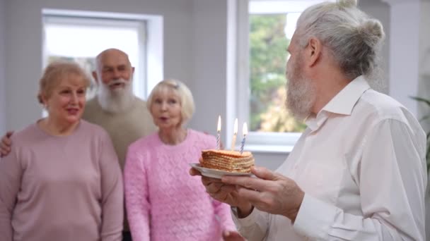 背景でお祝いの拍手を歌っている友人として誕生日ケーキの上にろうそくを吹いてハンサムな高齢者を笑顔 幸せな古いです白人男の肖像画上の右のパーティー — ストック動画