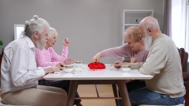 リビングルームでテーブルに座ってたくさん遊んで幸せな古い白人男性と女性のサイドビューの肖像画 テーブルゲームを屋内で楽しむ正の喜びの先輩の友人 — ストック動画