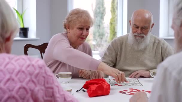 心情愉快的老朋友们坐在客厅里玩桌上彩票 积极快乐的白人老年男女在室内享受着游戏和休闲 — 图库视频影像