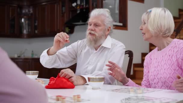 大胡子英俊的老人宣布与朋友在室内玩桌上彩票 积极的白人退休人员在客厅里享受着游戏 谈笑风生 — 图库视频影像