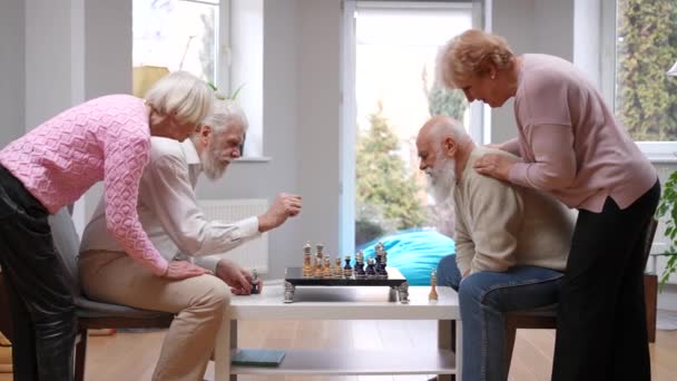 室内でチェスをする夫をサポートする正のシニア妻を表示します リビングルームでテーブルゲームを楽しむ男性を支持する幸せな白人女性 関係及び退職 — ストック動画