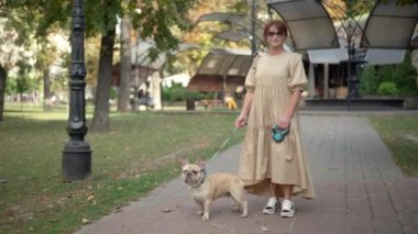 Şehir parkında safkan köpekle poz veren şık, olgun bir kadının geniş açılı portresi. Fransız Bulldog 'u ile kameraya bakan kendinden emin, güzel, beyaz, yaşlı bir kadın.