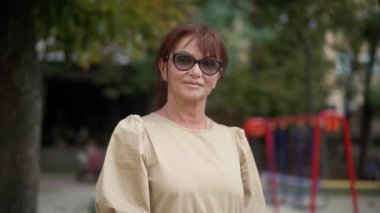 Şehir parkında kameraya bakarak duran gözlüklü ve elbiseli muhteşem, olgun, beyaz bir kadının orta boy portresi. Güzel, kendine güvenen bayan dışarıda poz veriyor.