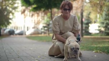 Yaşlı bir Fransız Bulldog 'u, bir gözü kapalı, yetişkin bir kadının evcil hayvanını okşadığı parkta. Geniş açılı, rahat, mutlu bir evcil hayvan ve güzel, beyaz bir ev sahibi.