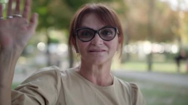 Gülümseyen, mutlu, olgun bir kadın. Gülümseyen, gülümseyen, kameraya bakan şehir parkında oturan. Kendine güvenen beyaz emeklinin portresi. Açık havada poz veriyor.